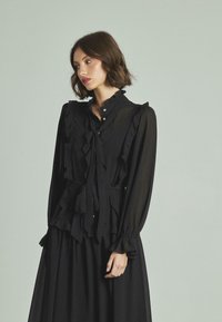 Vestido negro de manga larga con detalles de volantes, cuello alto y acentos de botones. Fabricado con un tejido ligero y suave, que ofrece una silueta fluida.