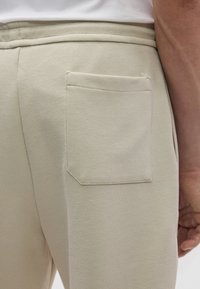 Beiget, teksturoidut housut, joissa näkyvä takatasku. Kangas vaikuttaa tukevalta ja siinä on hienovarainen kuvio. Käsi lepää sivulla.