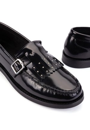 Mocassins en cuir noir brillant avec boucle argentée et franges cloutées sur fond blanc.