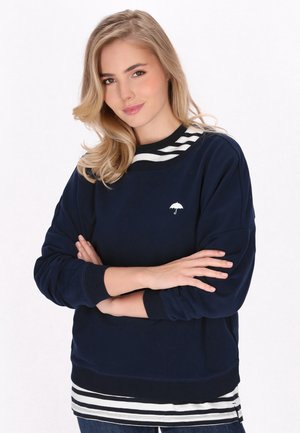 Laivaston sweatshirt, jossa on valkoinen sateenvarjologotulostus, raidallinen kaulus, joustavat hihansuut ja helma; kerroksittain raidallisen valkoisen ja laivaston sinisen paidan päällä.