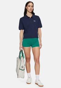 Marineblauwe poloshirt met korte mouwen en Fila-logo, gecombineerd met groene shorts. Model houdt een crèmekleurige tas met groene accenten vast en draagt witte sneakers.