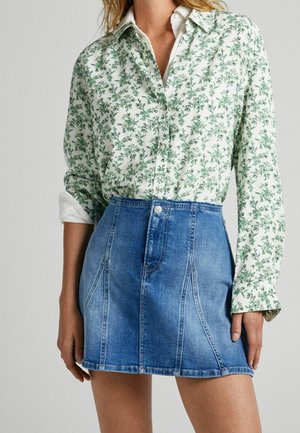 Woman wearing a white long-sleeve shirt with green floral print tucked into a blue high-waisted denim mini skirt.
