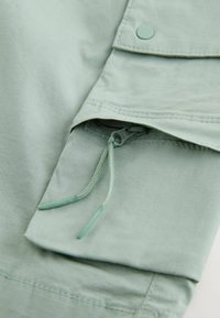 Détail de tissu vert clair montrant une poche zippée avec une languette de fermeture à cordon et un rabat sécurisé par un bouton-pression.