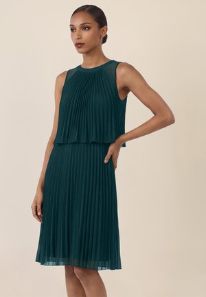 Frau trägt ein ärmelloses, dunkelblau-grünes, plissiertes knielanges Kleid mit einem mehrlagigen Oberteil und steht mit einer Hand in der Hüfte vor einem beigen Hintergrund.