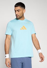 Γαλάζιο βαμβακερό μπλουζάκι με κοντά μανίκια, στρογγυλή λαιμόκοψη και πορτοκαλί λογότυπο Adidas στο στήθος. Φορεμένο με λευκά περιβραχιόνια.