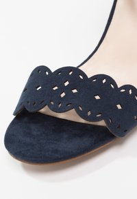 Gros plan d’une sandale en daim bleu marine avec une bride festonnée présentant des motifs découpés en forme de losange.
