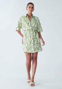 Kjole med korte ærmer og slå-om-design i cremefarve med grønt abstrakt mønster, der har bæltet talje og en løs pasform. Glat stof med blød tekstur.