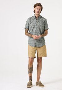 Kurzärmliges, grünes Hemd mit Knöpfen und einem Blättermuster, kombiniert mit beigen Shorts. Das Model steht mit zusammengelegten Händen und zeigt Tattoos sowie neutrale Schuhe.