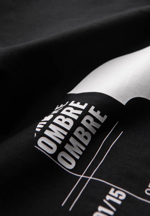 Tissu noir avec le texte en gras blanc "OMBRE" répété, formes géométriques partiellement visibles et fines lignes blanches sur une surface courbe.