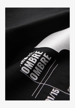 Tissu noir avec le texte en gras blanc "OMBRE" répété, formes géométriques partiellement visibles et fines lignes blanches sur une surface courbe.