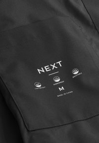 Prenda ligera negra con un bolsillo que presenta el logo "NEXT", iconos para propiedades impermeables y transpirables, y talla "M". Fabricado en China.