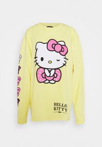 Camiseta amarilla de manga larga con un gran gráfico de Hello Kitty, un lazo rosa y detalles de lunares. Texto "HELLO KITTY" en el dobladillo.