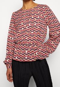 Langärmlige Bluse in Marineblau, mit floralem Muster in Rot-, Weiß- und Goldtönen, Samt der gerafften Taille und Rüschensäumen. Kombiniert mit gepunkteten schwarzen Hosen.