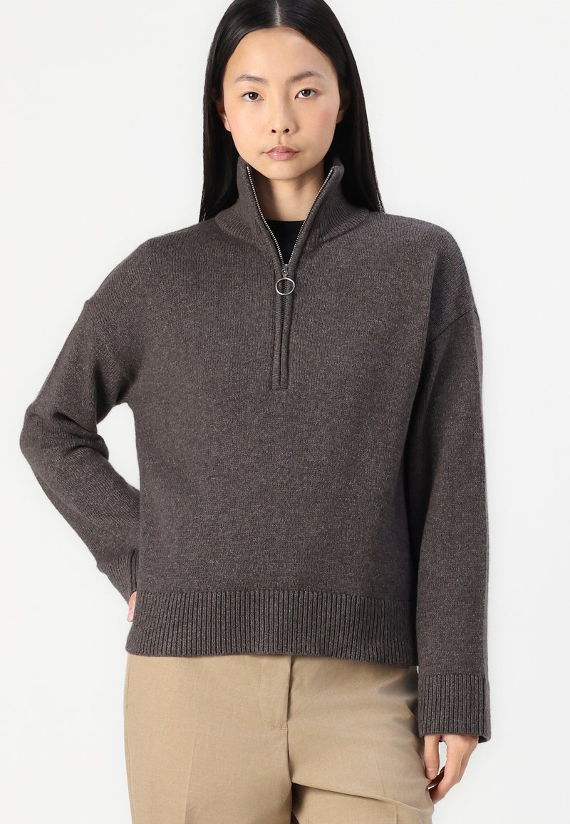 Brauner Strickpullover mit hohem Kragen, halbseitigem Reißverschluss und gerippten Bündchen und Saum. Getragen über beigen Hosen mit einer entspannten Silhouette.