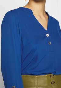 Blå långärmad blus med V-ringning, prydd med metallknappar och en slät textur. Bärs med ett grönt bälte.