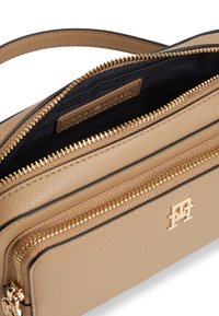 Μπεζ δερμάτινη τσάντα χειρός με χρυσά φερμουάρ, μικρό χρυσό λογότυπο στο μπροστινό μέρος και μαύρη εσωτερική επένδυση ορατή μέσα από το ανοιχτό φερμουάρ στο πάνω μέρος.