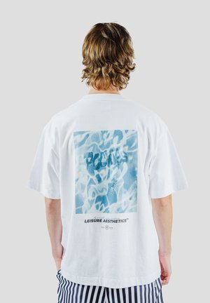 Person med krøllet brunt hår iført en hvid T-shirt med et blåt abstrakt vanddesign og teksten "LEISURE AESTHETICS" på ryggen.
