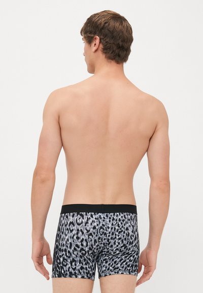 Mann som står med ryggen mot kameraet, iført grå og svart leopardmønstrede boxertruser med svart midjebånd.