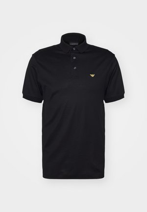 Črna polo majica iz gladkega bombažnega blaga, ki ima tri gumbe na ovratniku, kratke rokave in majhen zlat logotip na levem prsnem delu.
