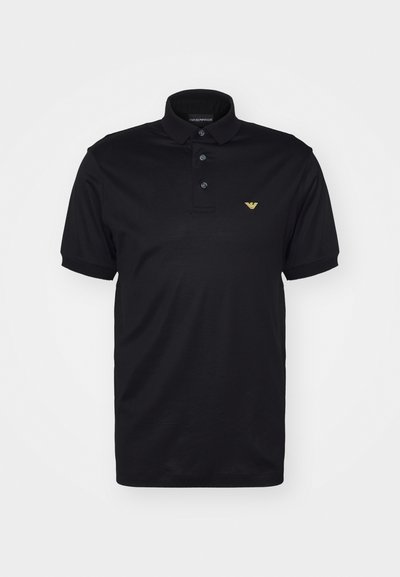 Črna polo majica iz gladkega bombažnega blaga, ki ima tri gumbe na ovratniku, kratke rokave in majhen zlat logotip na levem prsnem delu.