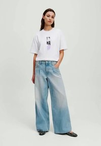 T-shirt branca com estampa gráfica, jeans azuis desbotados de perna larga e sapatos pretos de bico com pontos brancos. Design simples com um ajuste descontraído.