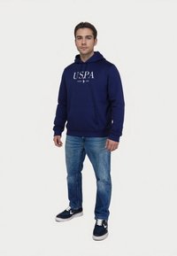 Tengerészkék kapucnis pulóver, az "USPA" logóval, elülső zsebbel és húzózsinórral. Kék farmernadrággal és tengerészkék sportcipővel párosítva, fehér kiegészítőkkel.