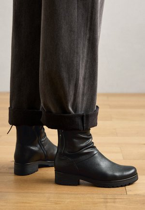 Schwarze Leder-Stiefeletten mit einem schlaffen Design, seitlichen Reißverschlüssen und Gummisohlen, kombiniert mit hochgekrempelten schwarzen Hosen. Hintergrund aus Holzboden.