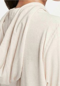 Ljusbeige hoodtröja med en avslappnad passform, som har en draperad huva och mjukt, texturerat tyg. Visar subtila sömnadsdetaljer på baksidan.