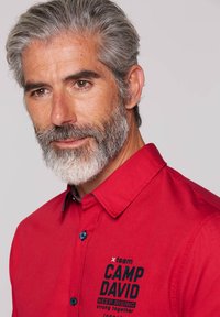 Rotes Button-Up-Hemd aus Stoff mit strukturierter Oberfläche. Verfügt über blaue Knöpfe und schwarzen Text auf der Vorderseite mit der Aufschrift "CAMP DAVID KEEP RISING."