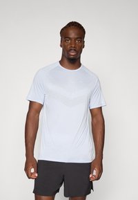 Cămașă sportivă scurtă de culoare albastră deschisă, cu un model texturat pe față, confecționată din material care absoarbe umezeala, asortată cu pantaloni scurți negri.
