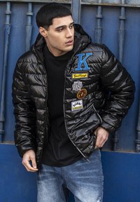 Chaqueta de plumas negra con capucha, adornada con múltiples parches coloridos y una textura brillante y acolchada. Se lleva sobre una camiseta negra y vaqueros.