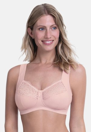 Blonde woman smiling, wearing a light pink bra with wide straps and lace detailing on the upper cups against a plain light background.