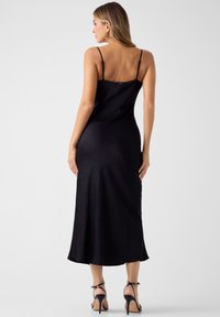 Μαύρο midi φόρεμα από σατέν με λεπτές τιράντες, με εφαρμοστή σιλουέτα. Ομαλή υφή και ελαφριά λάμψη, που φτάνει λίγο κάτω από το γόνατο.