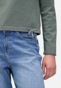 Grüner Sweatshirt mit strukturiertem Finish und Kordelzug am Saum, kombiniert mit hellblauen Jeans im klassischen Schnitt.