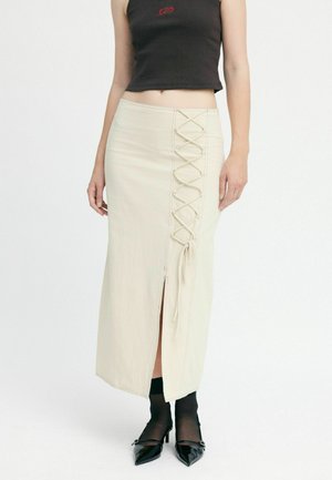 Beige midi skirt with a side slit, featuring lace-up detail along one side. Smooth texture, accompanied by black patent leather shoes.