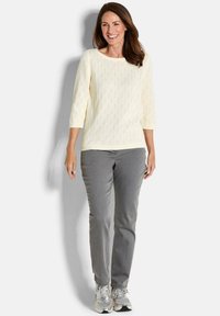 Cremefarbener gestrickter Pullover mit lockerer Passform und Rundhalsausschnitt, kombiniert mit grauen geraden Hosen und silbernen Sneakern.