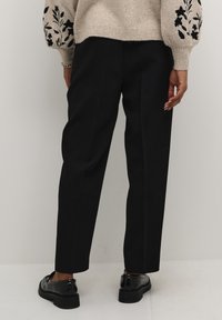 Pantalons tailleur noirs à coupe droite. Tissu lisse, taille haute et coutures latérales ; complétés par des chaussures noires brillantes.