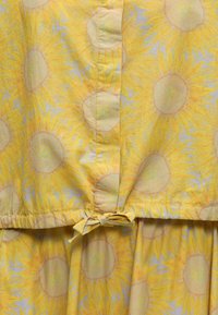 Tissu jaune avec de grands motifs de tournesols présentant un nœud noué en bas d'une ceinture froncée.