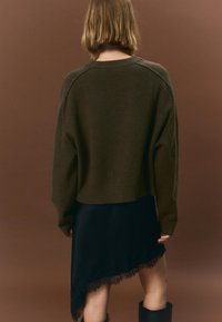 Pulover olive crop din material moale, asortat cu o rochie neagră cu tiv dantelat cu margini ondulate, pe un fundal maro.