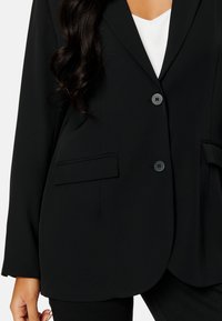 Czarna marynarka o dopasowanym kroju, z tradycyjnymi klapami, dwoma przednimi kieszeniami i zapięciem na jeden guzik. Miękki, strukturalny materiał o gładkiej teksturze.
