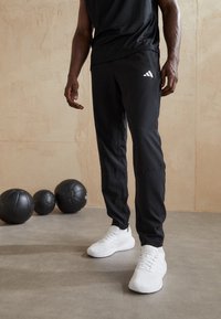 Svarta träningsbyxor med en avsmalnande design, med en vit Adidas-logga på vänster lår, i kombination med vita sneakers. Texturerat tyg.