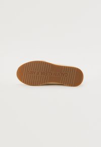 Podeszwa sneakersów Calvin Klein Jeans, z wzorzystą brązową gumową bazą, z rowkami i wytłoczonym logo marki. Gładkie, zaokrąglone krawędzie.