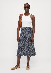 Un débardeur blanc associé à une jupe midi fleurie bleu marine. Lunettes de soleil et sac marron. Offre une silhouette fluide et une texture de tissu douce.