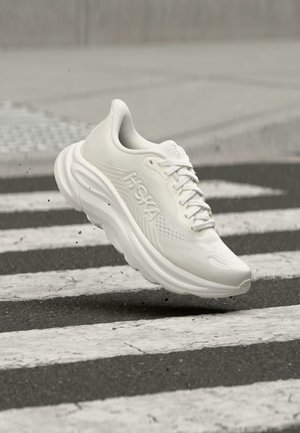 White Hoka running shoe hovering above a textured black and white pedestrian crossing on an asphalt street.
