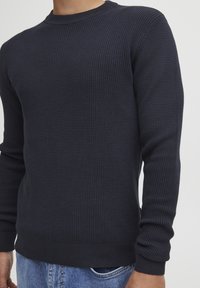Homme portant un pull à manches longues en maille gaufrée bleu marine et un jean bleu, visible du cou jusqu'aux hanches sur un fond uni.