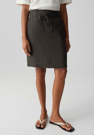 Vrouw draagt een donkergrijze knielange rok met een gebonden tailleband, een witte gebreide top en beige teenslippers, staand op een lichte achtergrond.