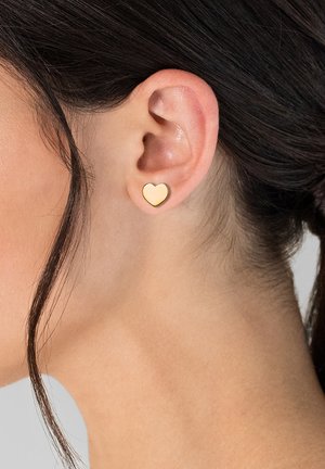 Goldfarbene herzförmige Steckerohrringe mit einer glatten, reflektierenden Oberfläche und schwarzer Umrandung, getragen an einem hellhäutigen Ohr. Einfaches und minimalistisches Design.