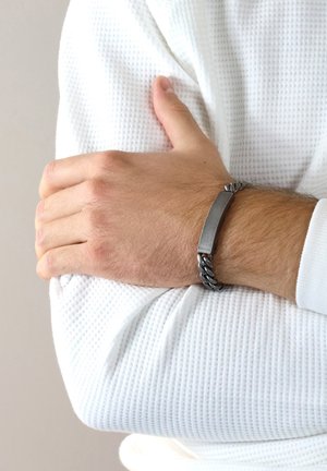 Metal bracelet with a flat, rectangular central piece, featuring a braided chain design in dark tones, worn on a wrist with textured white fabric.