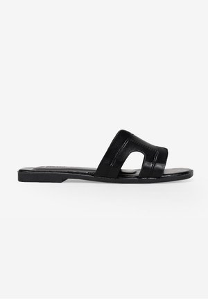 Sort flad slide sandal med bred rem med udskæringsdetaljer og firkantet tådesign på hvid baggrund.