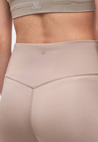 Gros plan sur le bas du dos et la taille d’une personne portant des leggings beige sans couture et un soutien-gorge de sport assorti avec de petits logos sur chaque vêtement.